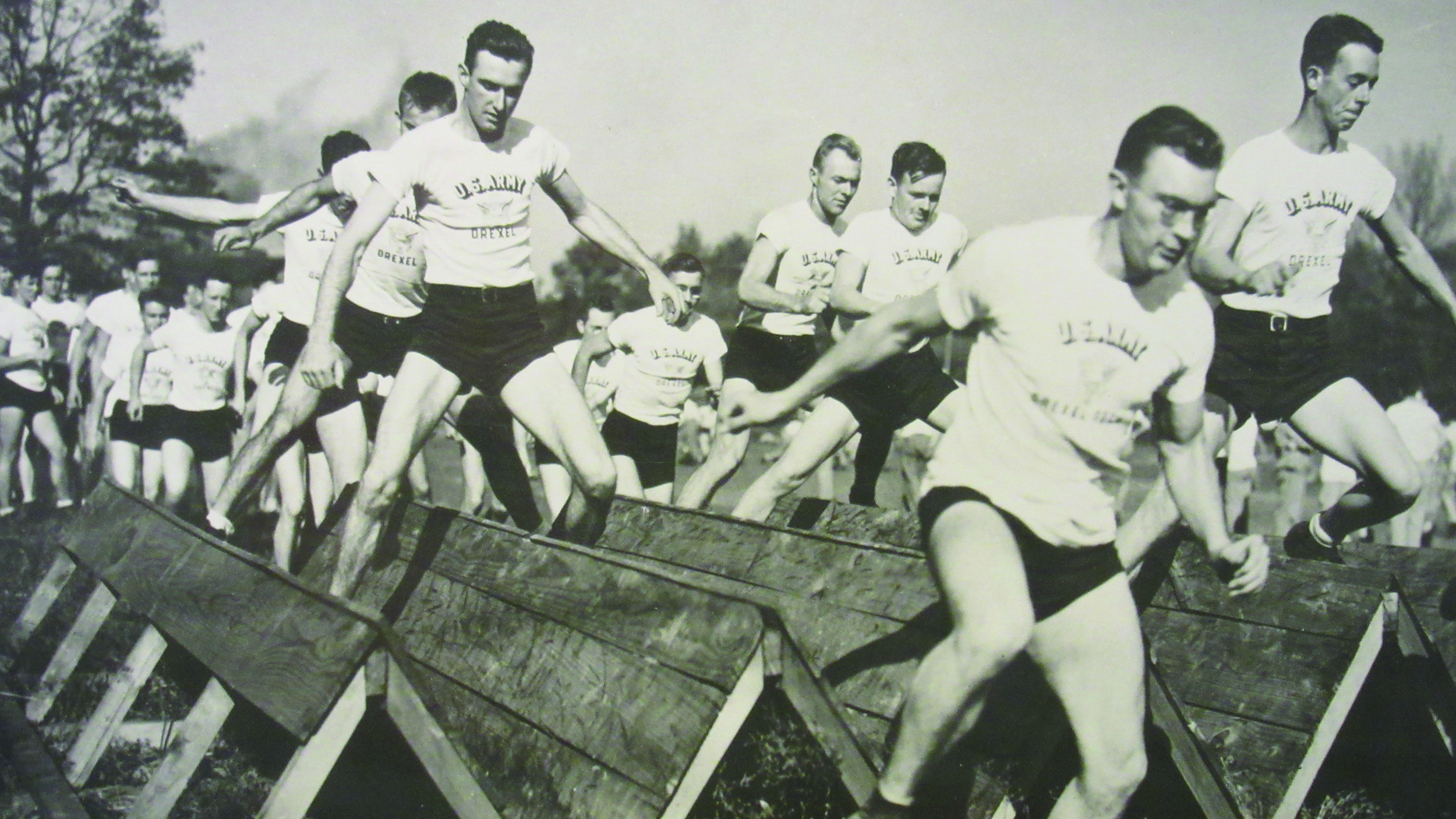 A black and white photo of a large group of U.S. Army cadets navigating an obstacle course, wearing shirts that say 'US Army, Drexel'.