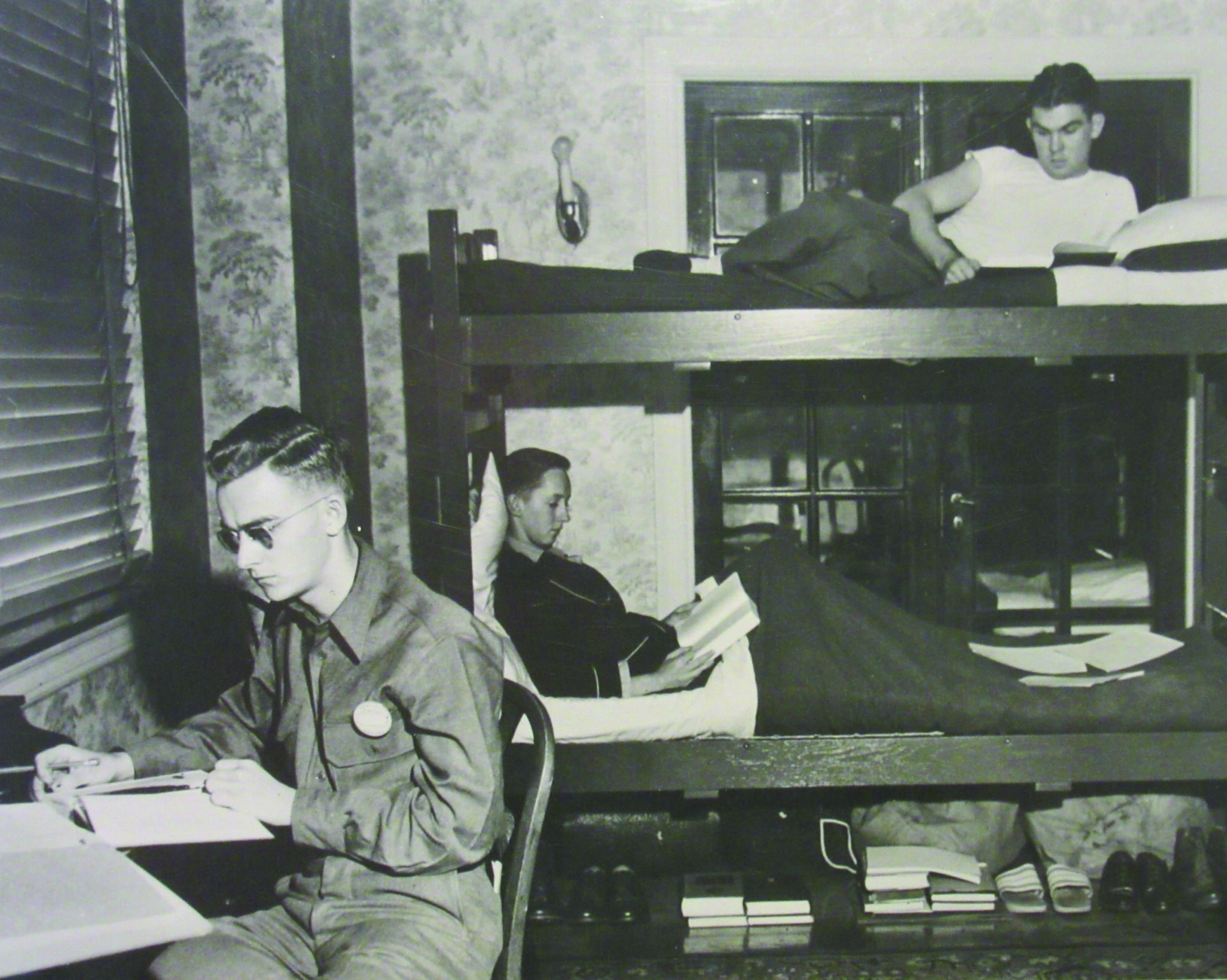A black-and-white photo of three cadets in a hotel bedroom, one studying at a desk and two reading in a bunk bed.
