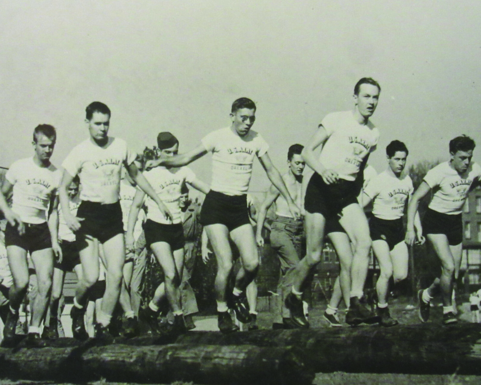 A black-and-white photo of a group of cadets running over large wooden telegraph poles placed sideways on the ground.