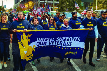 A group of people walk down a Philadelphia street carrying a banner that reads, "Drexel Supports Our Veterans."