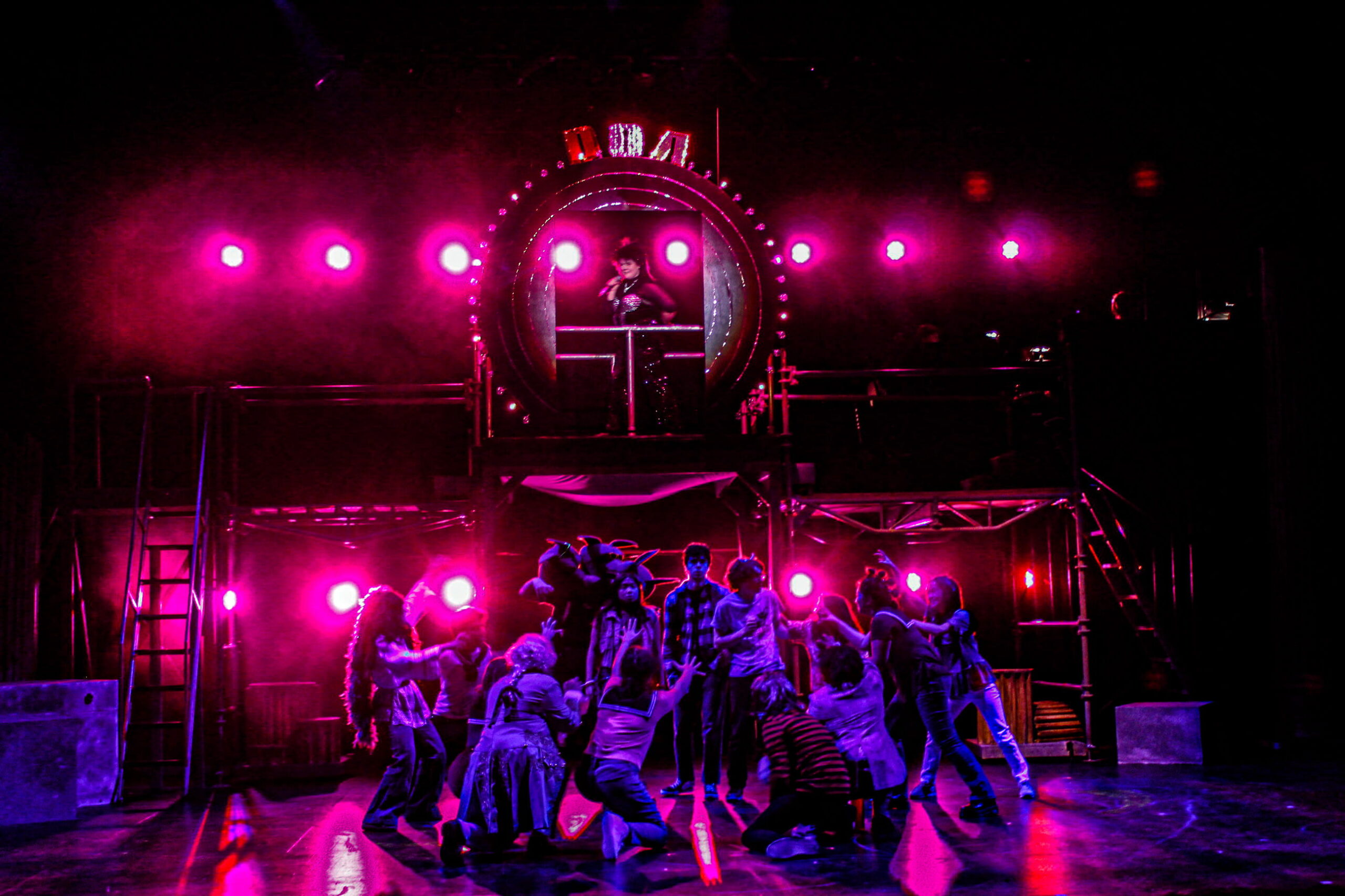 Student actors perform an ensemble musical scene on a pink-lit stage